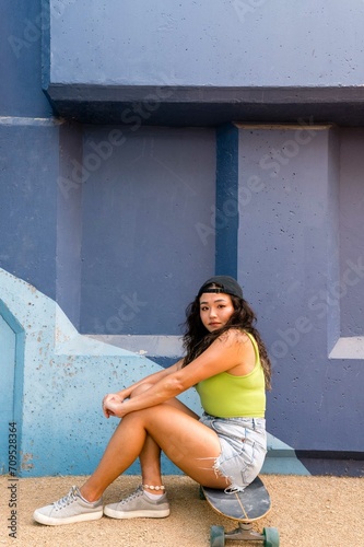 Asian woman sitting on her skateboard next to the blue wall © sashapritchard