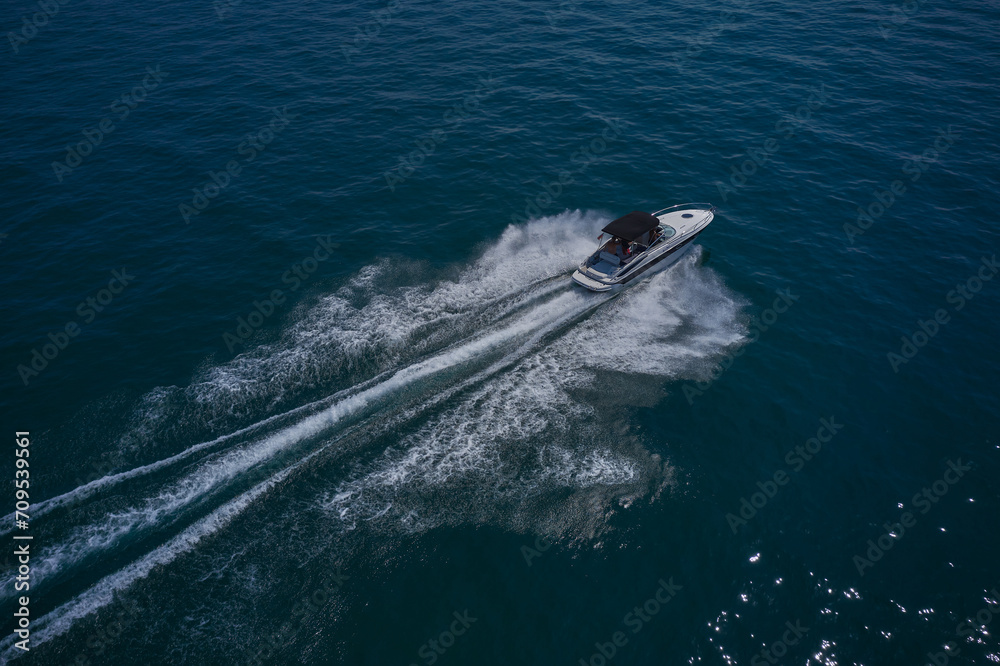 A large white boat with a dark awning moves quickly on blue water, moving diagonally, top view. A large boat with people moving on the water, aerial view.