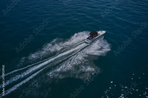 A large white boat with a dark awning moves quickly on blue water, moving diagonally, top view. A large boat with people moving on the water, aerial view.