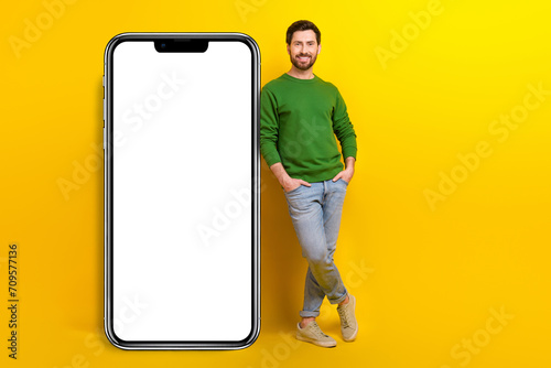 Full length photo of young businessman businessman posing near huge touchscreen phone application isolated on yellow color background