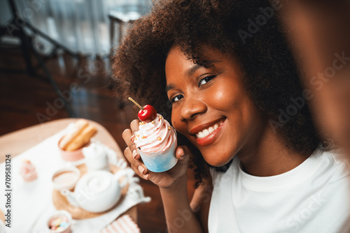 Beautiful young African blogger presenting piece of cupcake in concept special cuisine with selfie on smartphone. Content creating of social media with favorite sweets bakery dish. Tastemaker.