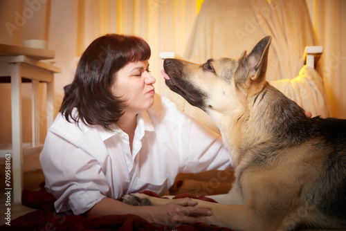 Adult mature woman with big shepherd dog in white shirt. Room with girl and calm cozy evening atmosphere with transparent curtains and soft warm light of lamps. Concept of love for animals and pets