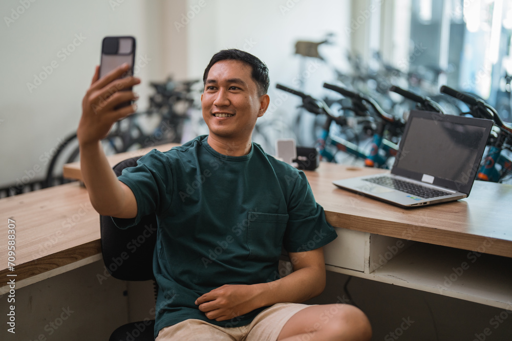 young man taking a selfie with his cell phone while working at his desk - obrazy, fototapety, plakaty 