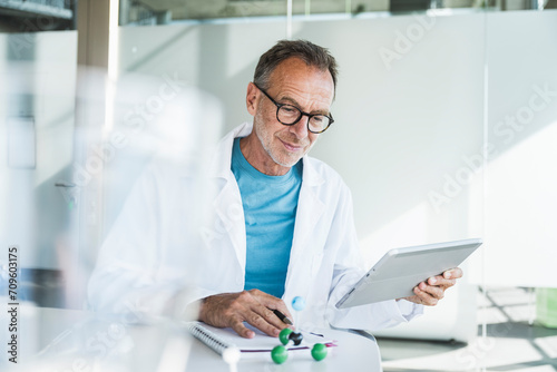Smiling senior doctor using tablet PC and sitting with note pad at desk photo