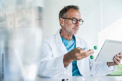 Man in lab coat holding molecular structure and using tablet PC at desk photo