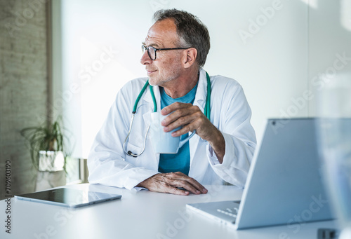 Contemplative senior doctor sitting with coffee cup at desk photo