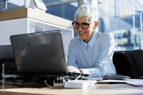 Smiling senior businesswoman using laptop sitting at desk in office photo