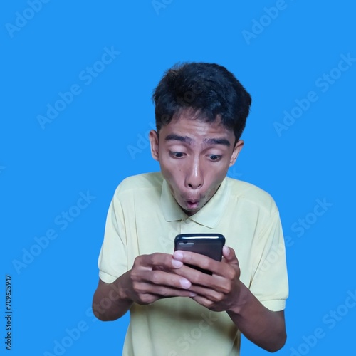 Young Asian man surprised looking at smart phone wearing yellow t-shirt, blue background.