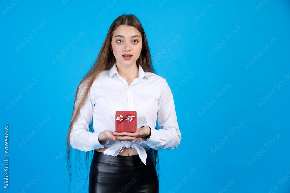 Amazed woman opening mouth from surprise, while getting gift prepared by friends. Astonished young girl holding small red box decorated with bow, isolated on blue background. Concept of emotions. Amazed woman opening mouth from surprise, while getting gift prepared by friends. Astonished young girl holding small red box decorated with bow, isolated on blue background. Concept of emotions.