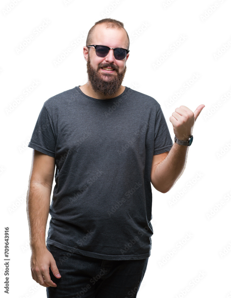 Young caucasian hipster man wearing sunglasses over isolated background smiling with happy face looking and pointing to the side with thumb up. Young caucasian hipster man wearing sunglasses over isolated background smiling with happy face looking and pointing to the side with thumb up.