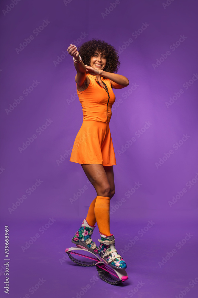 Sporty curly woman in orange outfit posing in kangaroo jumpers on studio background Sporty curly woman in orange outfit posing in kangaroo jumpers on studio background