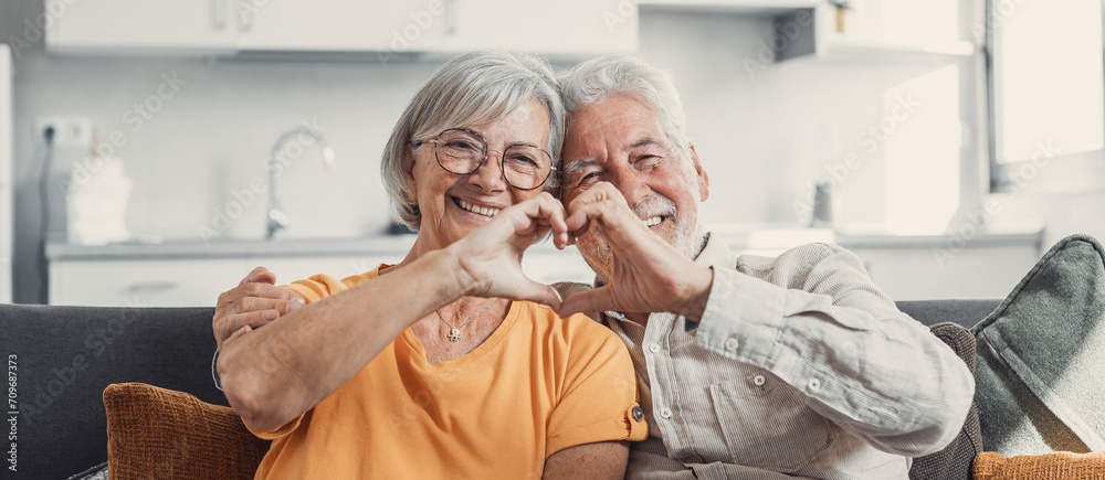 Close up portrait happy sincere middle aged elderly retired family couple making heart gesture with fingers, showing love or demonstrating sincere feelings together indoors, looking at camera.. - obrazy, fototapety, plakaty 