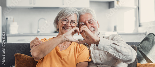Close up portrait happy sincere middle aged elderly retired family couple making heart gesture with fingers, showing love or demonstrating sincere feelings together indoors, looking at camera.. photo