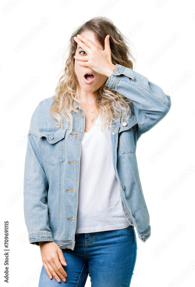Beautiful young blonde woman wearing denim jacket over isolated background peeking in shock covering face and eyes with hand, looking through fingers with embarrassed expression.