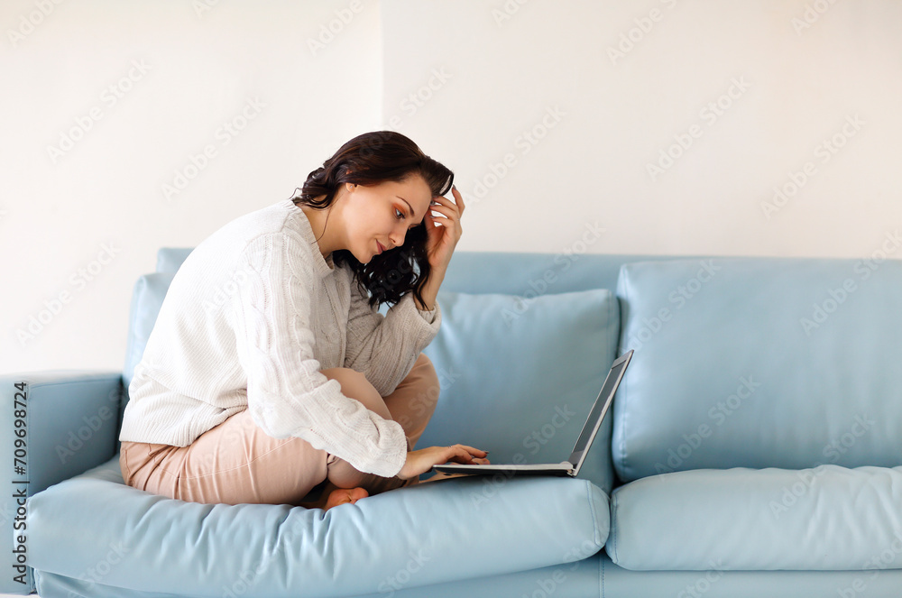 Young pensive woman in casual wear sitting with her legs on sofa with laptop. Freelance job