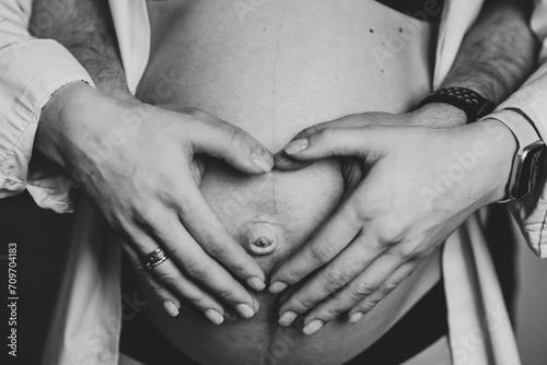 Pregnant woman and man hugging tummy at home closeup. Waiting baby. Husband hold belly of pregnant wife making symbol heart hands. Loving couple. Parenthood concept. Nine months. Black and white photo