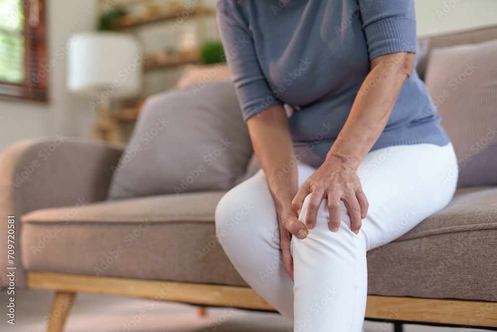 Old woman with knee pain sits on the sofa. Senile woman with body aches, leg pain sitting on a sofa in the living room stretching.