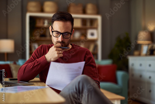 A focused male editor revising a draft work on a paper that he is holding while sitting in his home office. photo