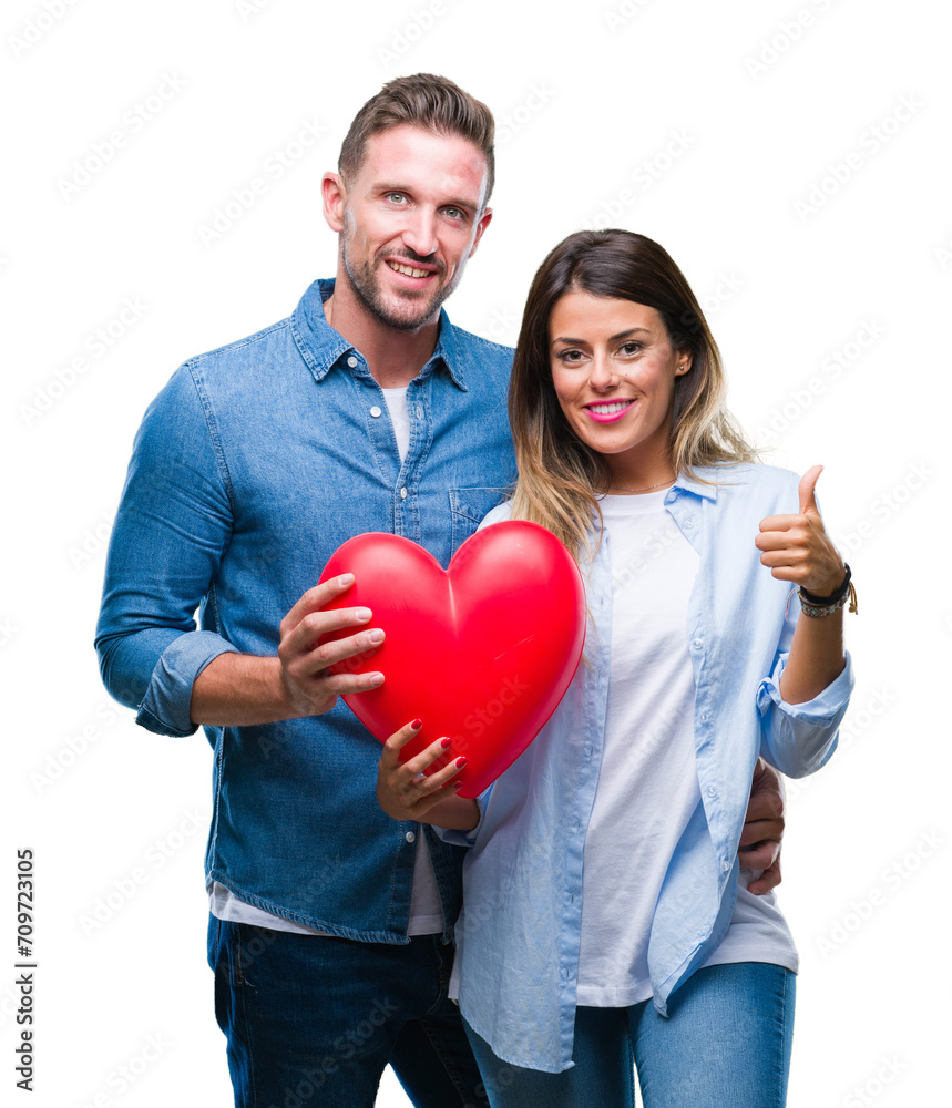 Young couple in love holding red heart over isolated background happy with big smile doing ok sign, thumb up with fingers, excellent sign