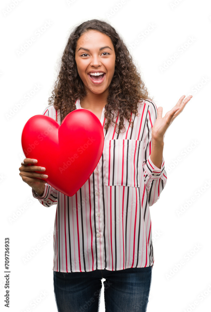 Young hispanic woman in love holding red heart very happy and excited, winner expression celebrating victory screaming with big smile and raised hands Young hispanic woman in love holding red heart very happy and excited, winner expression celebrating victory screaming with big smile and raised hands