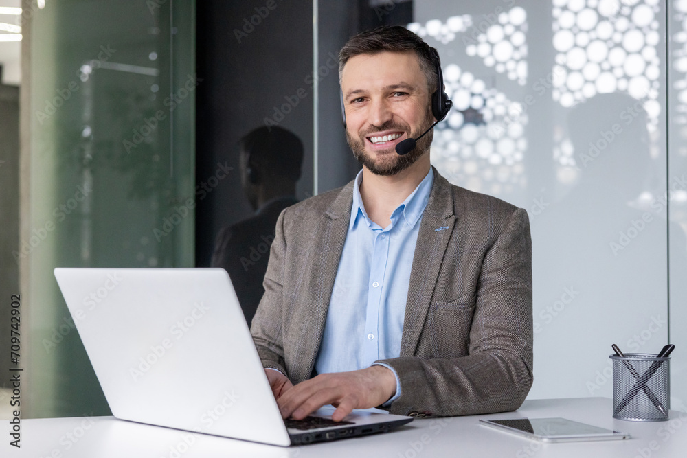 Smiling businessman with headset working on laptop in modern office setting, embodying professionalism and customer service.