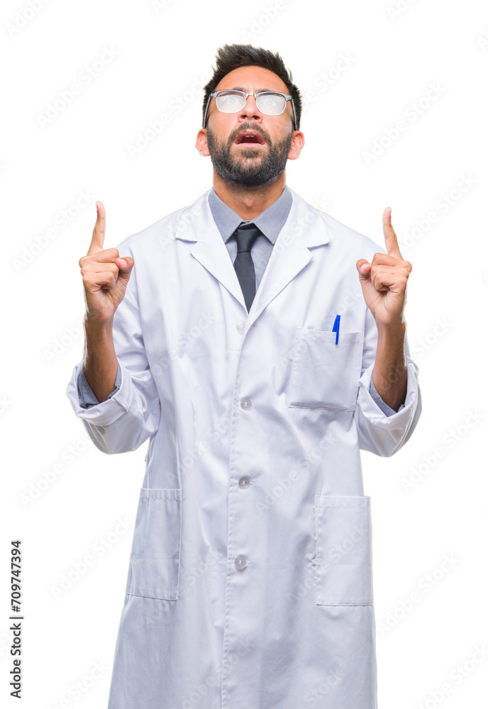 Adult hispanic scientist or doctor man wearing white coat over isolated background amazed and surprised looking up and pointing with fingers and raised arms. Adult hispanic scientist or doctor man wearing white coat over isolated background amazed and surprised looking up and pointing with fingers and raised arms.