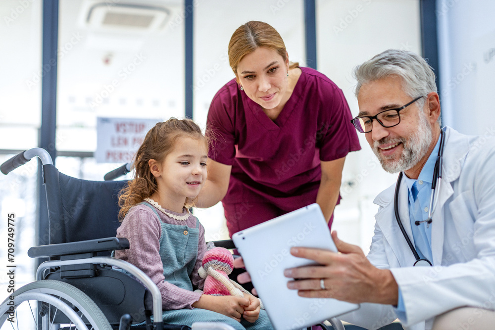 Fototapeta premium Friendly pediatrician showing someting on tablet to little patient in wheelchair. Cute preschool girl trusting her doctor and nurse in hospital.