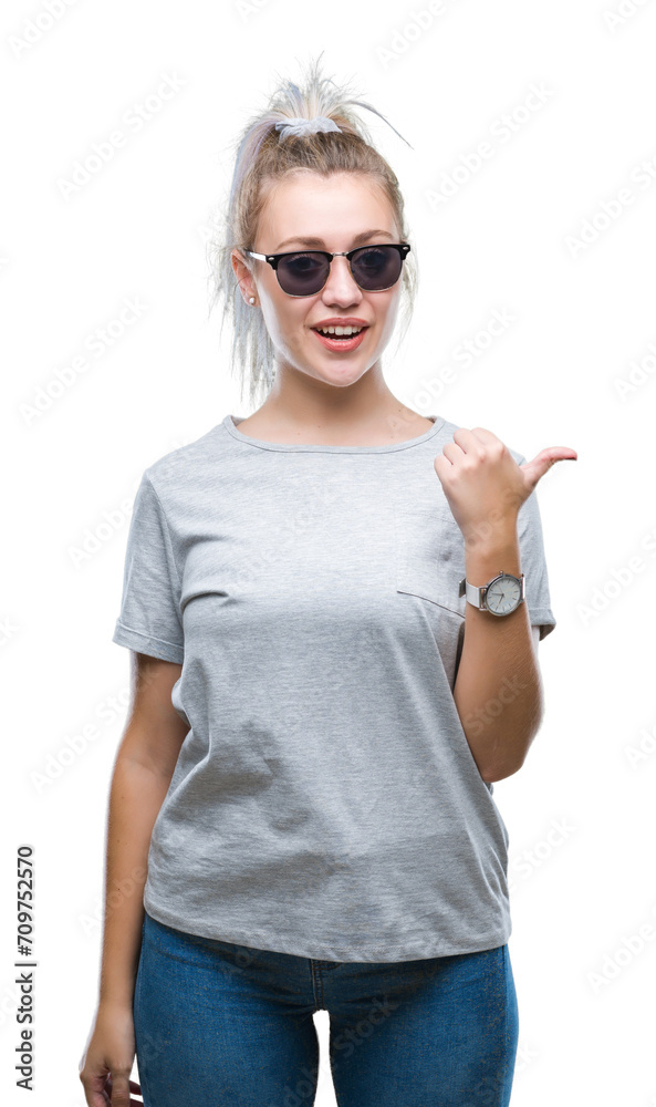 Young blonde woman wearing sunglasses over isolated background smiling with happy face looking and pointing to the side with thumb up.