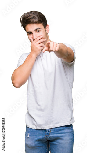 Young handsome man wearing white t-shirt over isolated background Laughing of you, pointing to the camera with finger hand over mouth, shame expression