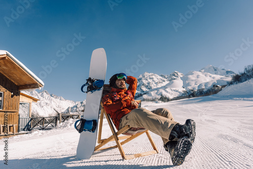 snowboarder sitting in sun lounger on sunny winter day, corduroy of prepared downhill slope at ski resort