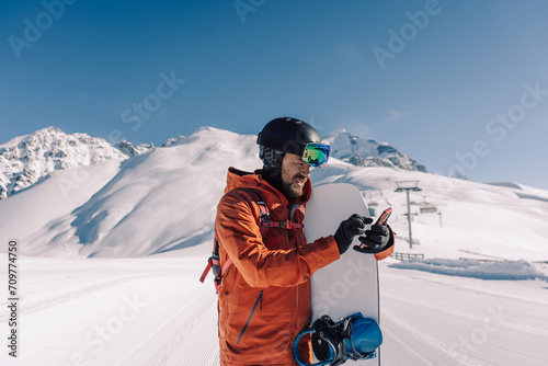 snowboarder holding snowboard and smartphone in his hands on prepared ski slope communicates by phone online and answers messages