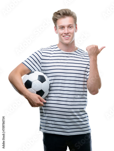 Young handsome blond man holding soccer ball pointing with hand and finger up with happy face smiling