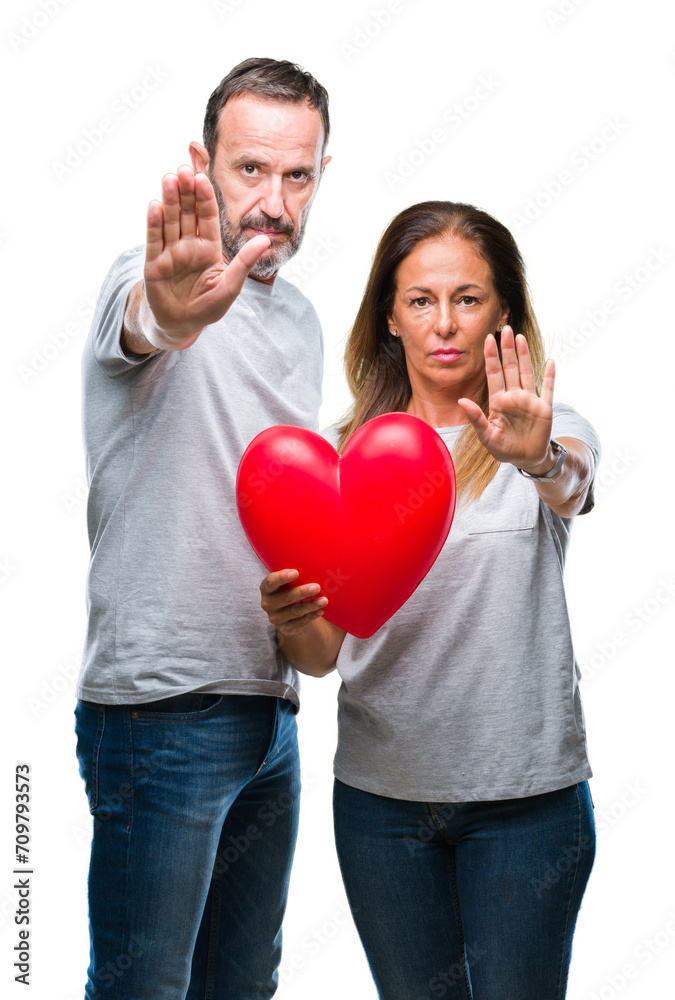 Middle age hispanic casual couple in love holding red heart over isolated background with open hand doing stop sign with serious and confident expression, defense gesture