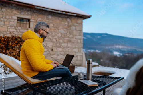 Mature man working from cozy cabin in mountains, sitting on terrace with laptop, enjoying cup of coffee. Concept of remote work from beautiful, peaceful location. Hygge at work. photo