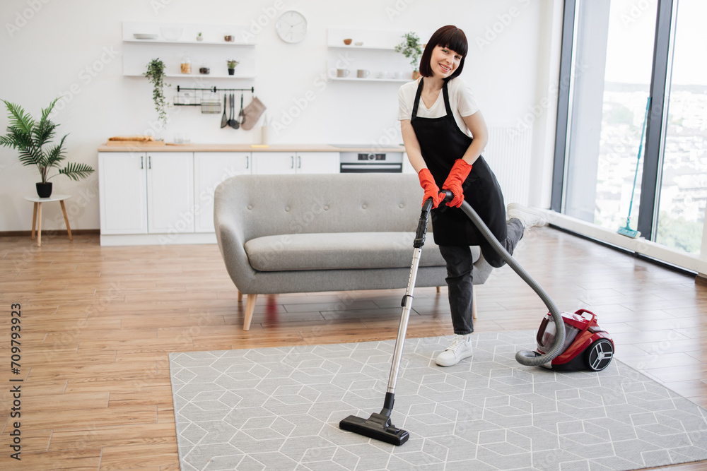 Professional female cleaner in apron actively cleans modern apartment and looking at camera. Positive woman in black uniform of cleaning service worker vacuuming carpet in stylish studio kitchen. Professional female cleaner in apron actively cleans modern apartment and looking at camera. Positive woman in black uniform of cleaning service worker vacuuming carpet in stylish studio kitchen.