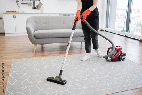 Cropped view of hands of woman in black uniform of cleaning service worker vacuuming carpet in stylish bright studio kitchen. Professional female cleaner in apron actively cleans modern apartment.