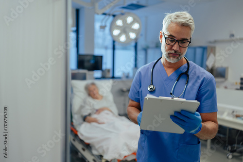 Portrait of handsome male doctor, patient in hospital bed behind. ER doctor examining senior patient, reading her medical test, lab results in clipboard. photo