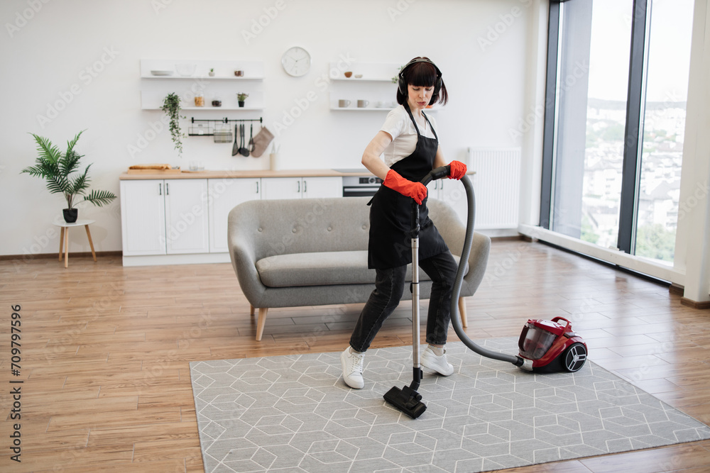 Fototapeta premium Positive young cleaners vacuuming rug of stylish modern studio kitchen. Cleaning service employee listens to music in headphones and dances while cleaning bright and spacious apartment.