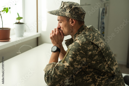 Thoughtful military man staring aside, holding palms by mouth, sitting on couch at home. Young soldier visiting psychologist, suffering from posttraumatic stress, closeup photo, copy space photo