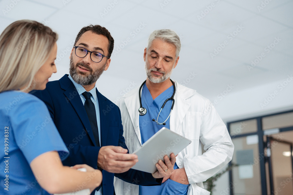 Fototapeta premium Pharmaceutical sales representative talking with doctors in medical building, presenting them new product on tablet, business talk. Hospital director consulting with healthcare staff.