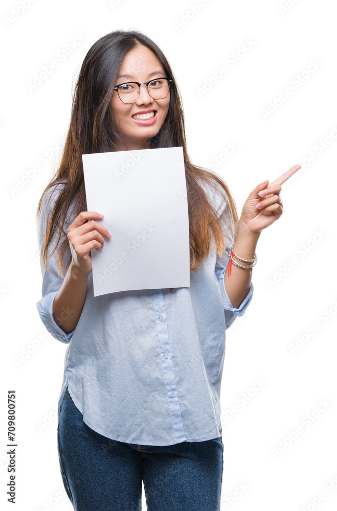 Young asian woman holding blank paper over isolated background very happy pointing with hand and finger to the side Young asian woman holding blank paper over isolated background very happy pointing with hand and finger to the side