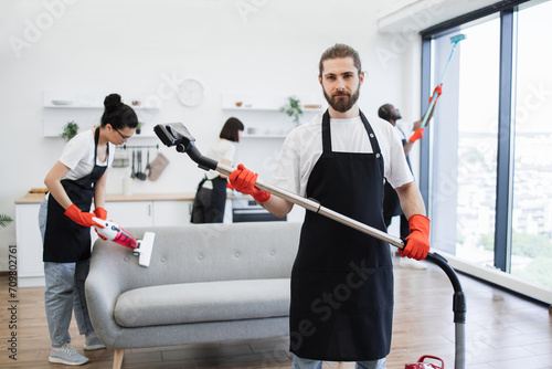 Portrait of smiling bearded cleaner from cleaning service holding wireless vacuum cleaner in hands, looking at camera, against background of multicultural colleagues cleaning bright, modern kitchen.