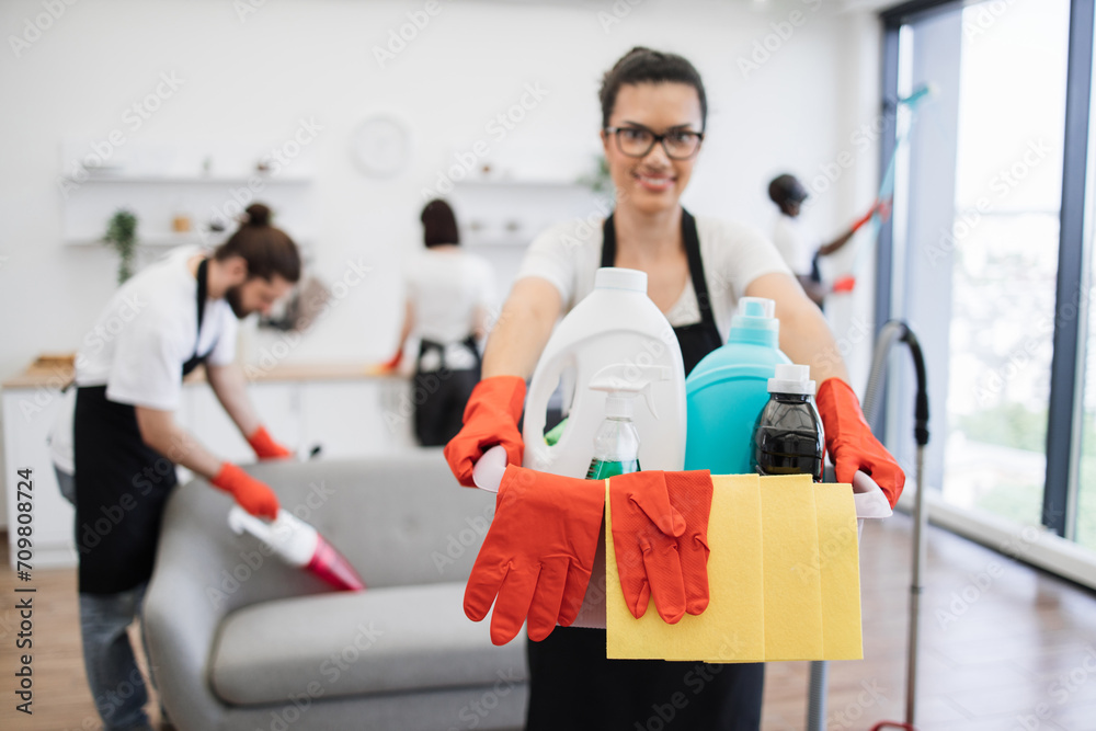 Focus on bucket for washing with detergents in hands of young African American woman professional cleaning worker bright kitchen studio background, copy space.