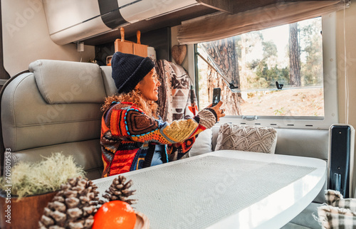 Woman wearing hat and pacwork sweater sitting in camper taking photos with cell phone looking out the window. Relaxation and leisure concept