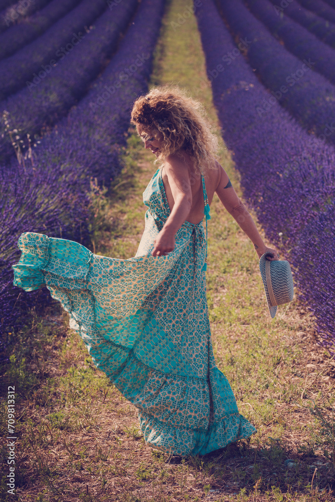 Back view of pretty lady in boho elegant blue dress walk in lavender fields wearing travel style hat and blonde curly nice long hair - concept or free woman in the outdoor nature adventure lifestyle Back view of pretty lady in boho elegant blue dress walk in lavender fields wearing travel style hat and blonde curly nice long hair - concept or free woman in the outdoor nature adventure lifestyle