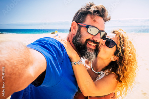 Adult couple of tourist take selfie picture at the beach with sand and blue ocean and sky in background. Hapy man and woman enjoying summer holiday vacation together. Travel lifestyle people photo