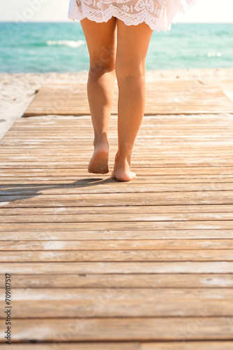 Back view of beautiful woman legs walking in summer on a wooden pier at the beach. Tropical summer holiday vacation people concept lifestyle. Tourist enjoying sun and blue water leisure activity