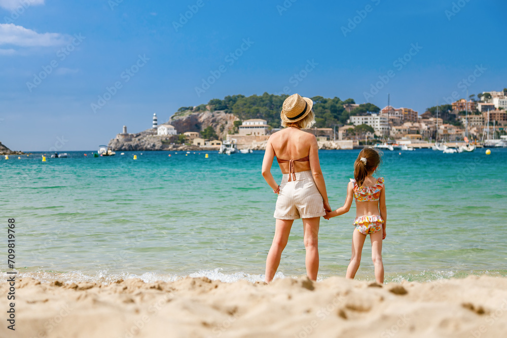 Woman and her little daughter in swimsuit standing together on the beach, watching at the Port de Soller bay in Mallorca
