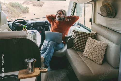 Digital nomad new modern job lifestyle with handsome adult man working and relaxing inside a camper van with beach and nature outside. Smart working free office concept eith laptop and connection