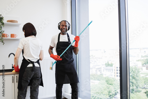 Team of professional cleaners on background of modern bright kitchen. African male worker washes panoramic windows, having fun while holding mop like guitar, listening to favorite song in headphones.
